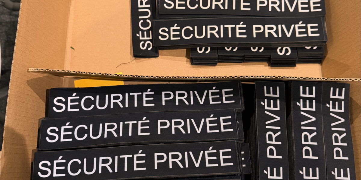 Carton rempli d’écussons rectangulaires noirs portant « SÉCURITÉ PRIVÉE » en blanc réfléchissant, rangés en piles